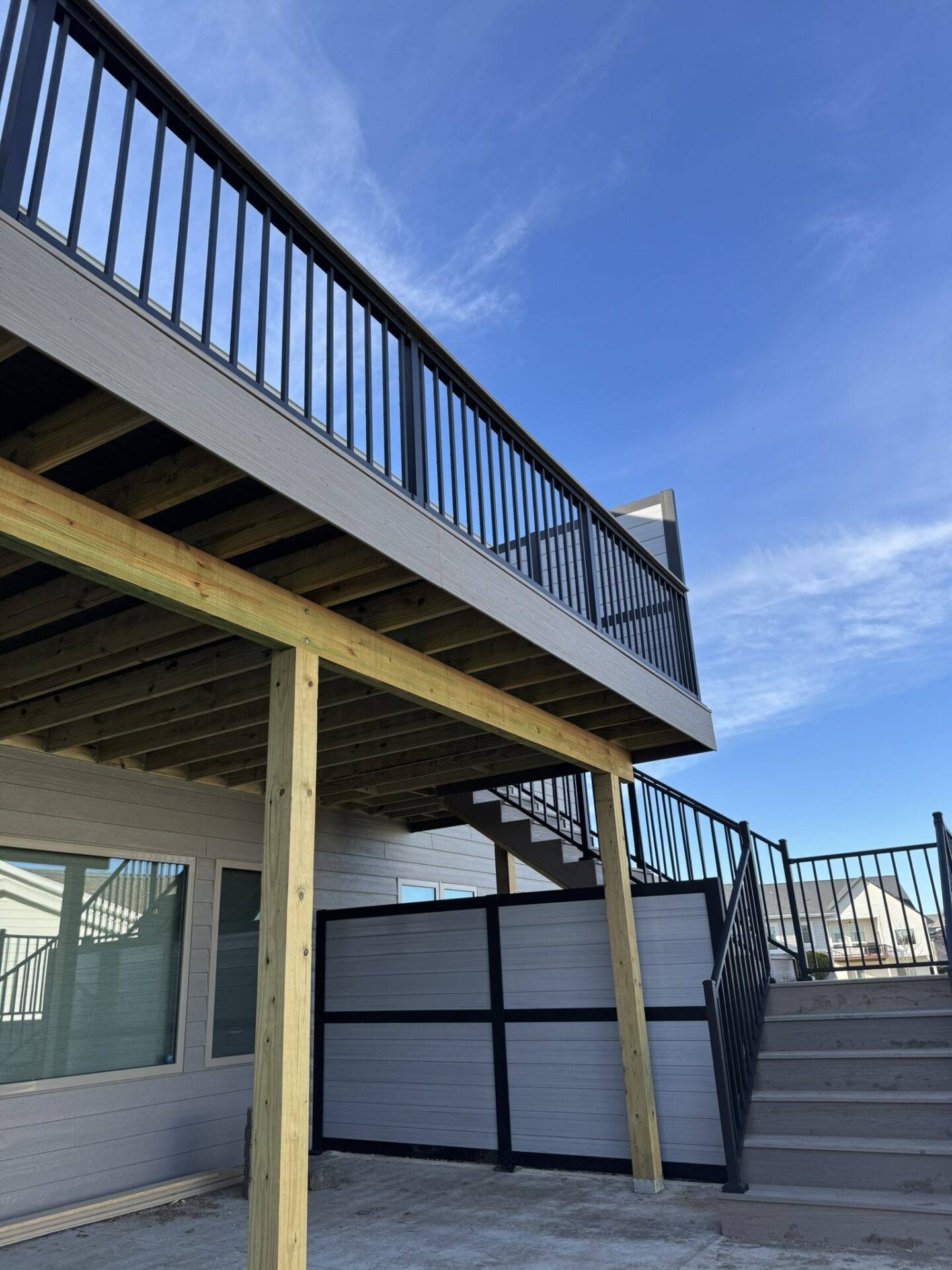 Underside of a new elevated composite deck showing treated posts, beams, joists, privacy screen and metal stairs on a Central Iowa home, deck building