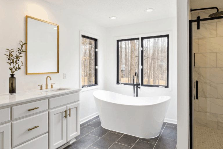 Busy Builders rendering of a bathroom remodel with freestanding soaking tub, black floor-mount tub filler, black frame windows, white shaker vanity, gold hardware, and dark slate tile flooring