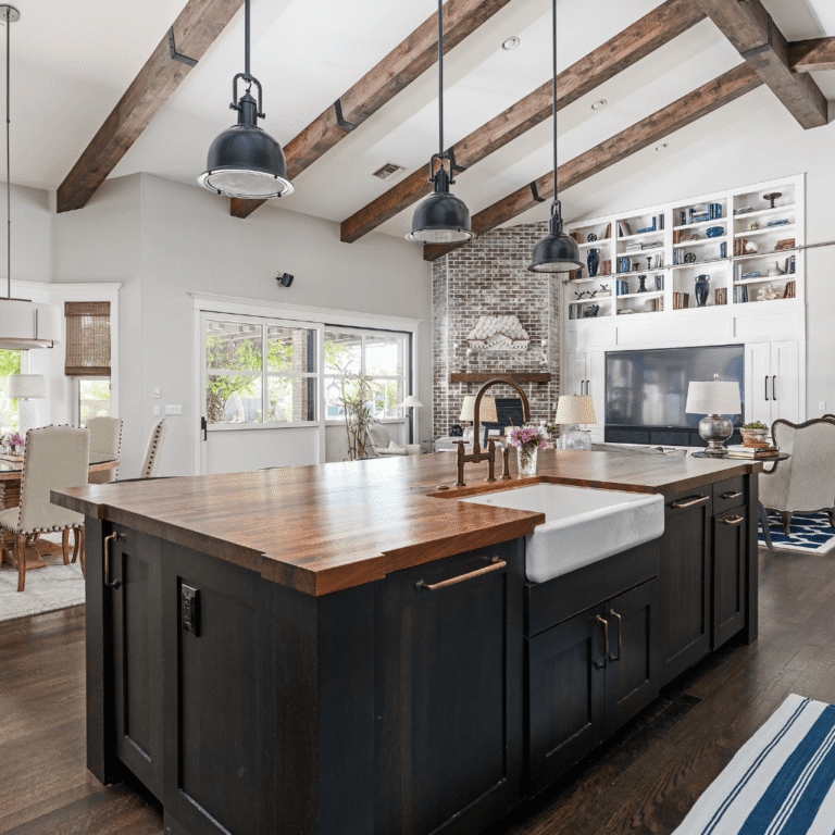 Busy Builders rendering of a home remodeling open plan kitchen with dark cabinetry, butcher block island, farmhouse sink, exposed wood beam ceiling, industrial pendant lights, and built-in shelving with brick fireplace