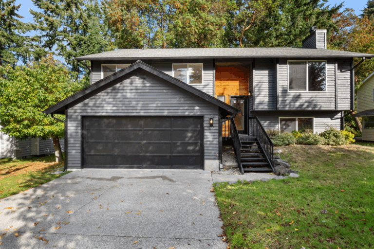 Split-level home exterior with dark charcoal gray siding, single-car attached garage with glass panel door, black metal stair railing, and natural wood front entry accent