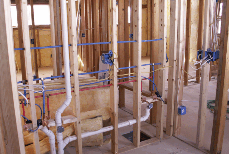 Plumbing and electrical rough-in with PVC drain pipes, blue and red PEX supply lines, and electrical wiring inside open wood stud framing