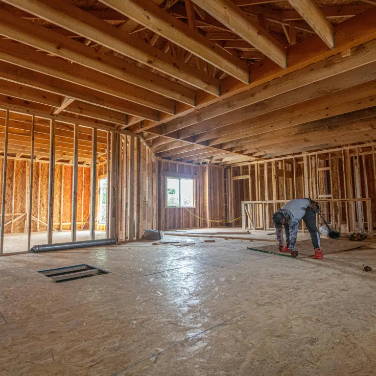 New home construction interior framing with open plan vaulted ceiling, OSB subfloor, exterior wall sheathing, and crew working on framing layout