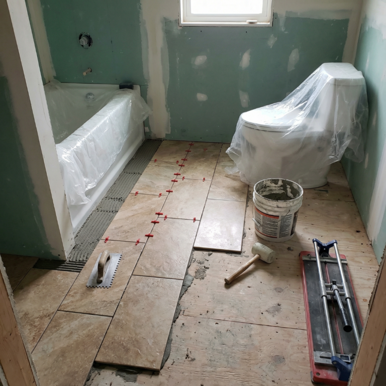 Bathroom remodeling tile installation in progress with floor tiles, spacers, thinset mortar, notched trowel, and protected toilet and bathtub on cement board walls